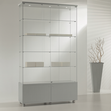 The Premier Lite 12.22M Tall Display Cabinet features three glass shelves and a lockable design with a grey base and two doors. To the right, a tall grey planter with bare branches enhances the minimal, bright decor.