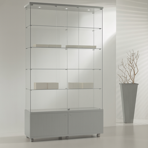The Premier Lite 12.22M Tall Display Cabinet features three glass shelves and a lockable design with a grey base and two doors. To the right, a tall grey planter with bare branches enhances the minimal, bright decor.
