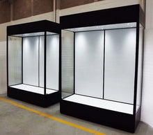 Two Double Mannequin Showcase display cabinets with lockable hinged doors and braked castors stand against a wall in a well-lit indoor space with a concrete floor.