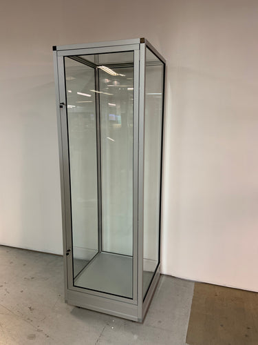 The Single Mannequin Showcase with Glass Top, a tall lockable display cabinet with a metal frame, stands on a concrete floor against a white wall. Its transparent glass door and interior space are well-lit by overhead lights.
