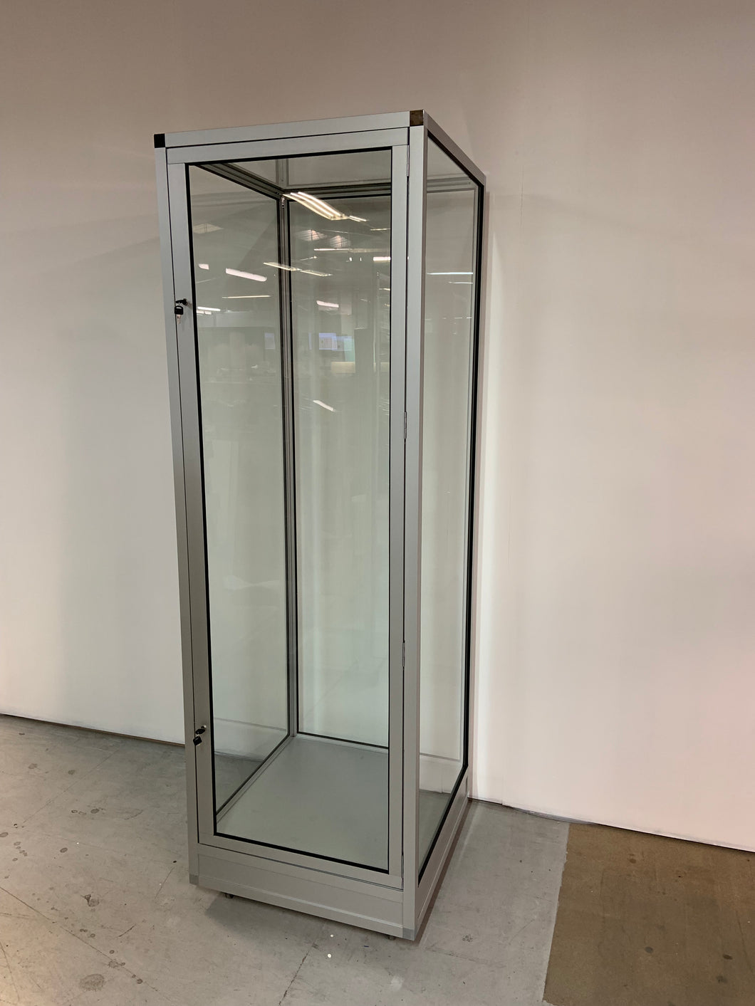 The Single Mannequin Showcase with Glass Top, a tall lockable display cabinet with a metal frame, stands on a concrete floor against a white wall. Its transparent glass door and interior space are well-lit by overhead lights.