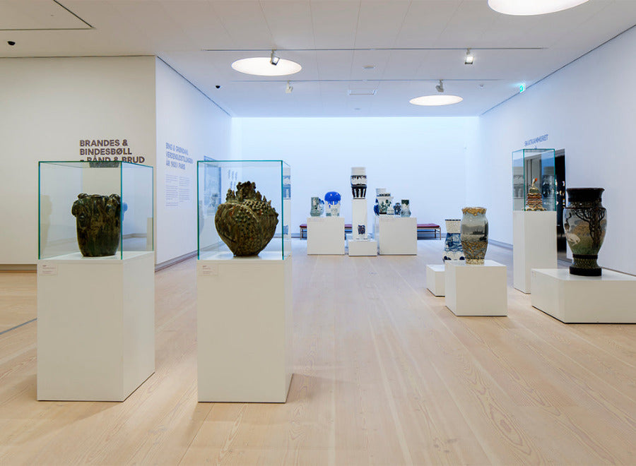 A modern museum gallery with light wood floors, white walls, and glass display cases showcasing various ceramic vases and pottery pieces on white pedestals. The space is brightly lit with overhead lights.