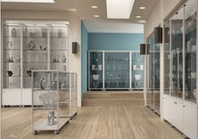 A modern museum gallery features Fusion Plus 5PLP Jewellery Glass Display Cases with lockable doors, displaying ceramics and decorative objects on a light wood floor, illuminated by spotlights against a blue accent wall.