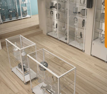 The Fusion Plus 5PLP Jewellery Glass Display Case with lockable doors displays silver bowls and sculptures, elegantly arranged in a bright, modern room with light wood floors and white walls.