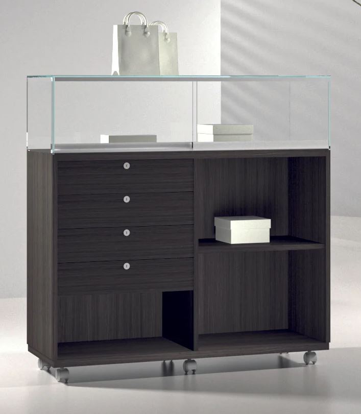 The Premier Lite 12.120B Wood Display Counter features a dark wood veneer base, glass top, lockable drawers, open shelves, display boxes inside, and wheels. A silver handbag and two boxes are displayed on and inside it in a brightly lit room.