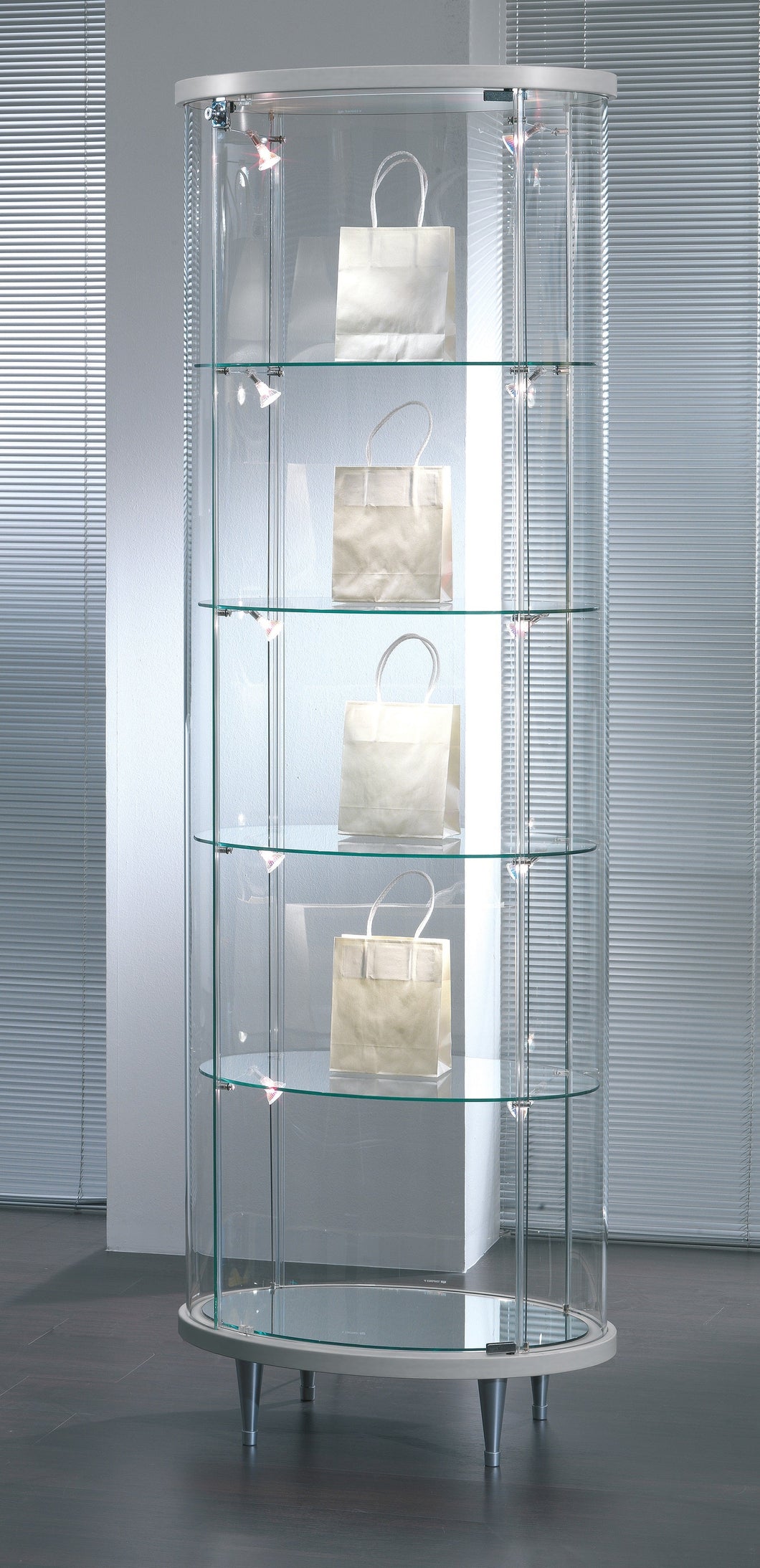 The Elegance Lite 203/OP Oval Display Showcase features a sleek design with transparent tempered glass shelves, showcasing stylish paper bags inside. This stunning glass display cabinet is perfect for highlighting your prized possessions while adding elegance to any space.